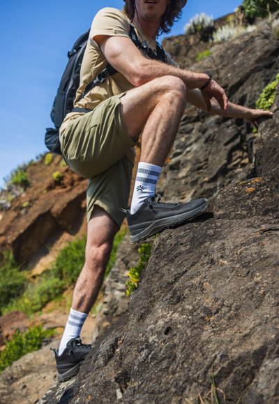 Grå vandringsskor med texturerade sulor, ihop med vita strumpor med blå ränder. Personen har på sig khakishorts och en beige tröja.