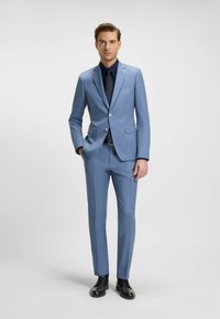 Completo blu chiaro realizzato in tessuto liscio, con giacca a un solo petto, pantaloni slim-fit e una leggera lucentezza. Abbinato a una camicia scura e scarpe.