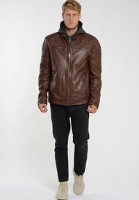 Brun læderjakke med lynlås foran, ribbede skulderdetaljer og et gråt hætte-skjold under, parret med sorte jeans og beige sneakers.