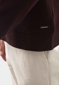 Mørk bordeaux ribbet sweatshirt med et metal logo mærke, parret med cremelagte fløjlsbukser med subtile vertikale striber.