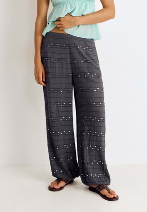 Mujer vistiendo pantalones oscuros grises y sueltos con rayas horizontales de lentejuelas, una camisa sin mangas de color verde claro y sandalias marrones, de pie frente a un fondo blanco.