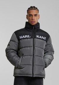 Grå pufferjakke med sorte skuldre, med lynlås lukning, høj krave og hvid "KARL KANI" logo på forsiden. Struktureret, polstret design.
