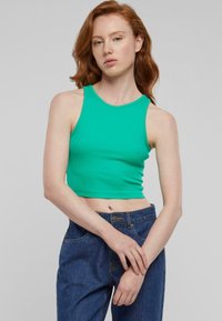 Zelený crop top vyrobený z elastického materiálu, s okrúhlym výstrihom a bezrukávovým dizajnom. V kombinácii s modrými džínsami.