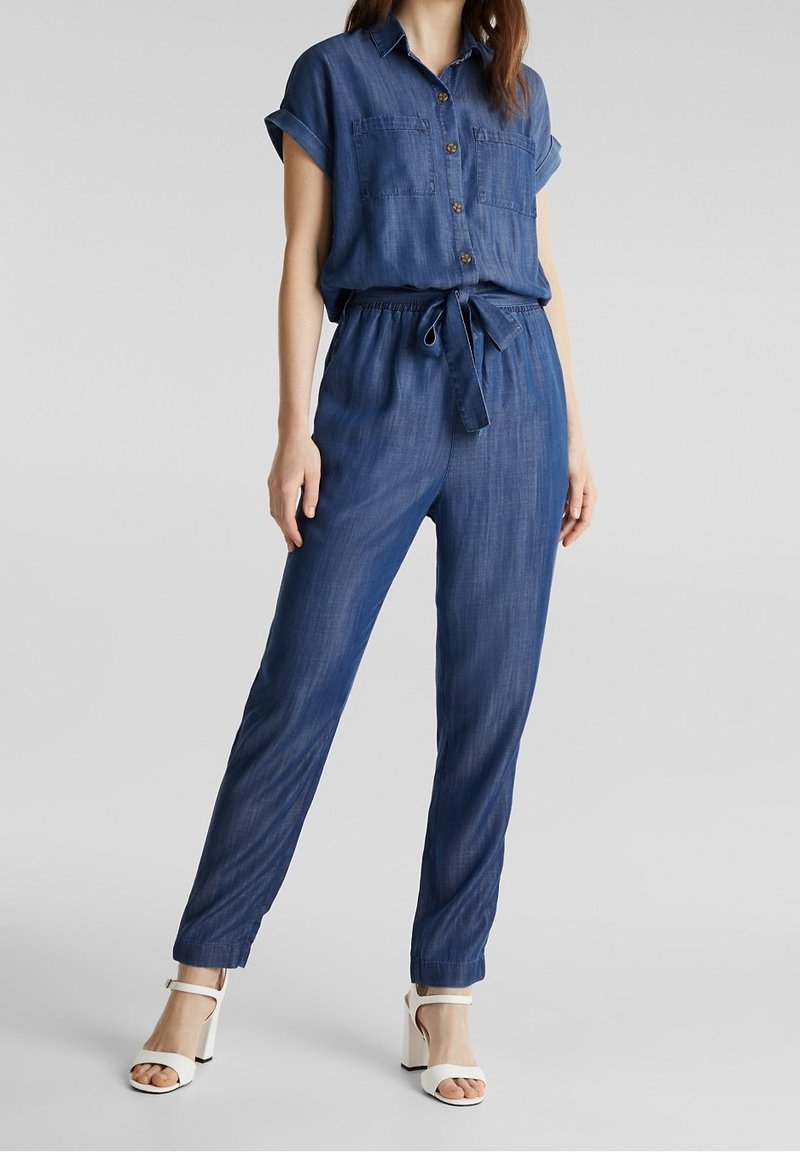 Combinaison en denim à manches courtes, à boutons sur le devant, avec deux poches poitrine, taille ceinturée et jambes effilées. Couleur : bleu profond. Tissu texturé.