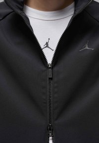 Chaqueta negra con cremallera fabricada en un tejido suave, que presenta un destacado logo de Jordan en el pecho y una camiseta blanca debajo con el mismo logo.