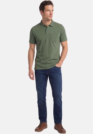 Mann trägt ein olivgrünes Poloshirt mit Brusttasche, dunkelblaue Jeans und braune Ledersneaker, steht mit einer Hand in der Tasche.