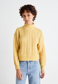 Pulover galben din tricotaj cu un guler înalt și ribatat, cu mâneci lărgi, asortat cu blugi de culoare albastru deschis. Textura este groasă și călduroasă.