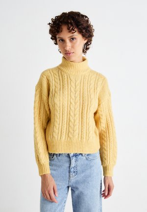 Maglione giallo a trecce con un alto colletto a coste e maniche morbide, abbinato a jeans azzurri chiari. La texture è spessa e calda.