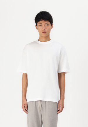 T-shirt blanc à manches courtes en coton, avec un col rond et une coupe décontractée. Le tissu semble lisse avec un design épuré.