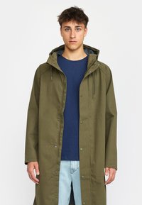 Olivgrüner Parka mit Kapuze, Druckknöpfen und Kordelzügen. Getragen über einem blauen Shirt, kombiniert mit hellblauen Jeans. Glatte Textur.