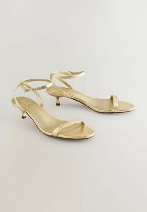 Kuldne rihmadega sandaalid, millel on peen pahkluu rihm, madal kitten konts ja minimalistlik disain. Sile metallic viimistlus ja avatud varvas.