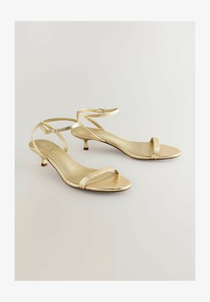 Kuldne rihmadega sandaalid, millel on peen pahkluu rihm, madal kitten konts ja minimalistlik disain. Sile metallic viimistlus ja avatud varvas.