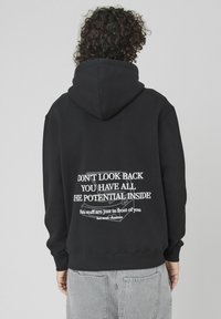 Hoodie noir en mélange de coton, avec un grand motif de texte blanc au dos comprenant des citations motivantes et une texture douce et lisse.