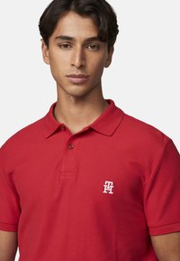 Camisa polo roja con una tela texturizada, que presenta un cuello y una tapeta de dos botones. Logo bordado en blanco en el lado izquierdo del pecho.