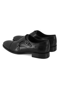 Schwarze Lederschuhe mit runder Zehenpartie, doppelten Mönchsschnallen und dekorativen Brogue-Details an der Vorderseite und den Seiten.