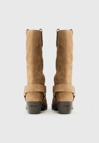 Par čez mid-kalv škornjev iz brušenega semiša rjave barve z zaponkami in črnimi petami na blok, prikazanih od zadaj na enostavni ozadju.