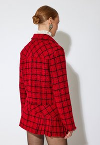 Blazer in tweed a quadri rossi con controlli neri, tessuto texturizzato, bordi sfilacciati e vestibilità su misura. Presenta un colletto a revers e spalle strutturate.