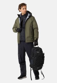 Abbigliamento da outdoor per uomo con giacca impermeabile verde scuro, gilet imbottito nero, pantaloni neri, guanti e un grande zaino nero.