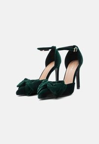 Zöld bársony magassarkú cipők hegyes orral, elöl egy nagy masnis díszítéssel és csatolható bokaszíjjal.