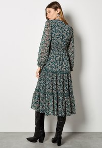 Vestido de mangas largas con volantes en azul verdoso y un patrón floral. Presenta una cintura fruncida y mangas translúcidas. Combinado con botas negras hasta la rodilla.