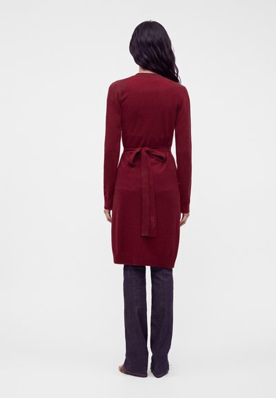 Femme vue de dos portant un cardigan bordeaux longueur genoux noué à la taille avec un pantalon droit foncé sur fond uni.