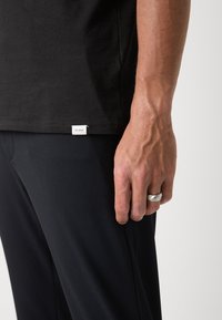 Tricou negru din bumbac cu o etichetă albă a brandului la tiv. Mâna arată un inel argintiu, asociat cu pantaloni negri. Textură netedă, design casual.