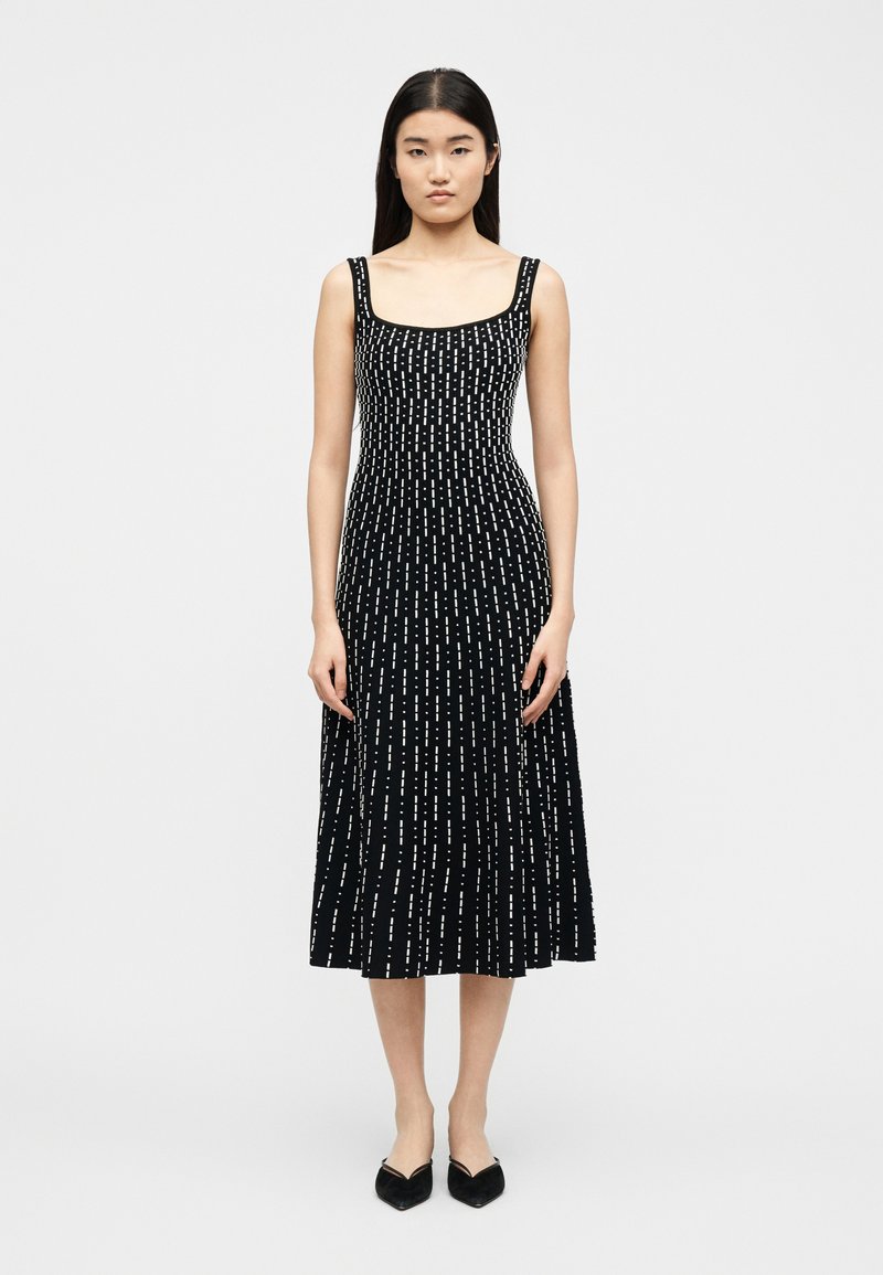 Femme portant une robe midi noire avec des motifs de tirets verticaux blancs, un décolleté carré, des bretelles fines et des ballerines noires pointues, debout devant un fond blanc.