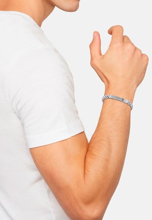 Pulsera en tono plateado con un diseño trenzado y un acento de metal plano, llevada en una muñeca con un tono de piel claro. Camisa blanca lisa de fondo.