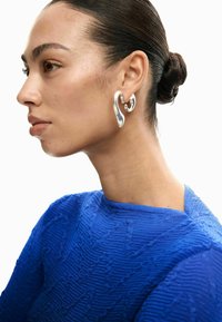 Frau mit zurückgekämmten Haaren zu einem Dutt gebunden, trägt ein strukturiertes blaues Oberteil und einen großen, verdrehten silbernen Ohrring am linken Ohr, blickt nach links.