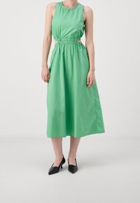 Vestido sin mangas verde con cintura fruncida y detalle recortado, hecho de una tela ligera. Llevado con tacones negros de punta afilada.