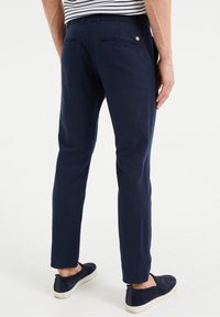 Man i marinblå chinos och marinblå slip-on skor, stående med ryggen mot kameran mot vit bakgrund.