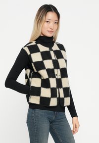 Gilet in pile checkato bianco e nero con scollo rotondo, chiusura frontale con bottoni e tasche laterali, indossato sopra un dolcevita nero.