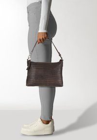 Braune Lederhandtasche mit strukturierten horizontalen Streifen, Reißverschluss und abnehmbarem Trageriemen, gehalten von einer Person, die graue Hosen und weiße Sneakers trägt.