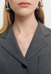 Kobieta z długimi blond włosami, nosząca duże srebrne kolczyki w formie kół i szarą marynarkę zapinaną pod szyją, usta zamknięte, oczy niewidoczne.