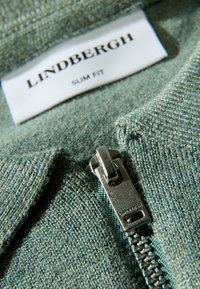 Zielona dzianina o dopasowanym kroju, z srebrnym zamkiem błyskawicznym i metką marki "LINDENBERGH". Zbliżenie na teksturę.