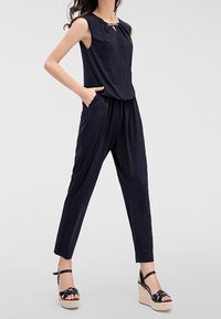 Marinblå jumpsuit med ärmlös design, rund halsringning och nyckelhålsdetalj. Har sidofickor och avsmalnande ben, kombinerad med svarta kilklackar.