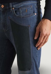 Jean en denim bleu foncé avec un panneau texturé noir sur la cuisse, agrémenté de rivets en cuivre et d'un contour cousu. Main reposant sur le côté.