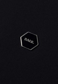 Pixie neagră hexagonală cu litere argintii în relief "BALR." pe un fundal texturat negru. Marginea metalică evidențiază forma.