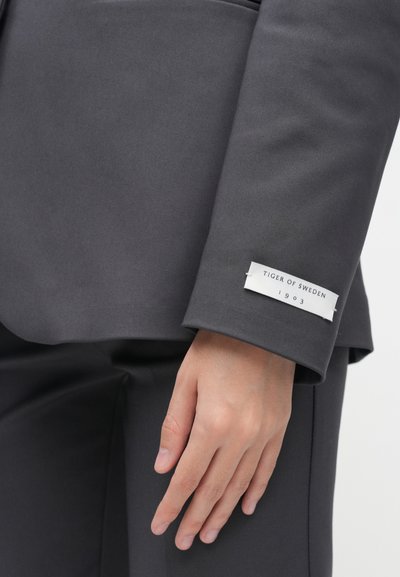Γκρι σακάκι με λεία υφή. Ο κύβος διαθέτει μία λευκή �ετικέτα με τις λέξεις "TIGER OF SWEDEN 1903" σε μαύρο κείμενο. Χέρι ακουμπισμένο πάνω στο σακάκι.