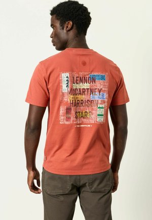 Mann trägt rostfarbenes T-Shirt mit buntem Grafikdesign und Text, der auf der Rückseite The Beatles' Lennon, McCartney, Harrison, Starr auflistet.