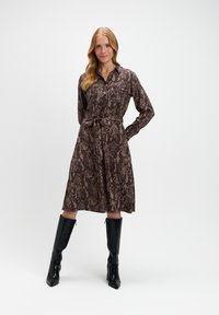 Brązowa sukienka koszulowa w wężowy wzór z kołnierzykiem, długimi rękawami i wiązanym w pasie. Połączona z czarnymi, wysokimi kozakami na słupku.