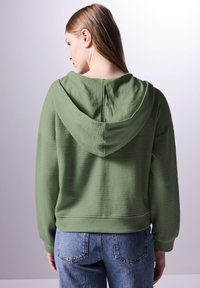 Felpa verde strutturata con un grande cappuccio, vestibilità ampia e maniche lunghe, abbinata a jeans in denim, fotografata da dietro.