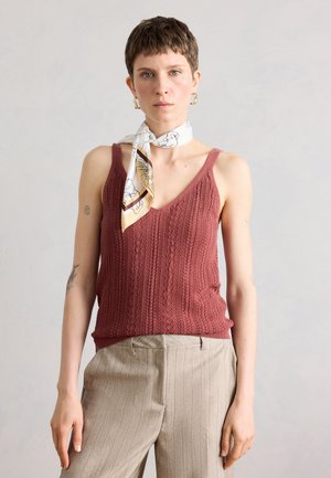 Osoba s krátkymi vlasmi, ktorá má na sebe vínový pletený tielko, béžové nohavice s tenkými pruhmi, biely vzorovaný šál na krku a zlaté krúžkové náušnice na jednoliatom pozadí.