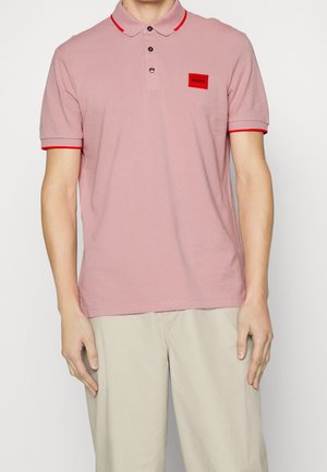 Man draagt een lichtroze poloshirt met rode bies en een rode Hugo-logo applicatie, gecombineerd met lichtbeige broek, staand tegen een witte achtergrond.