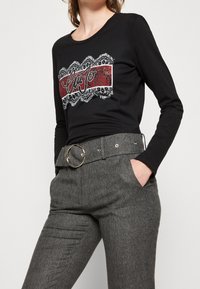 Schwarzes Langarmshirt mit einem roten und weißen Grafikdesign sowie Spitze. Kombiniert mit grauen Hahnentritthosen und einer großen silbernen Gürtelschnalle.