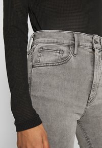 Jeans gris de cintura alta con una textura sutil, diseño de cinco bolsillos y cremallera, combinados con una blusa negra de manga larga con textura.