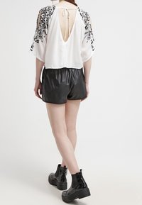 Blouse blanche avec broderie florale noire, découpe profonde dans le dos, associée à un short en similicuir noir et des bottines noires épaisses.