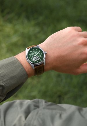 Rokas pulkstenis ar zaļu ciparnīcu, redzamām pārnesumiem, brūnu ādas siksniņu un sudraba korpusu, fonā nesaistīts zaļš fons.