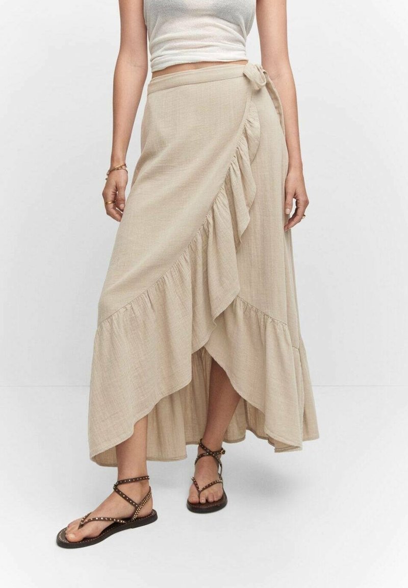 Femme portant une jupe portefeuille beige à volants, associée à un top court blanc et des sandales marron à lanières cloutées.