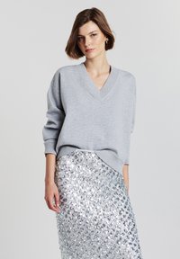 Donna con i capelli corti castani, che indossa un maglione grigio chiaro a V e una gonna di paillettes argentate, in piedi contro uno sfondo chiaro e semplice.